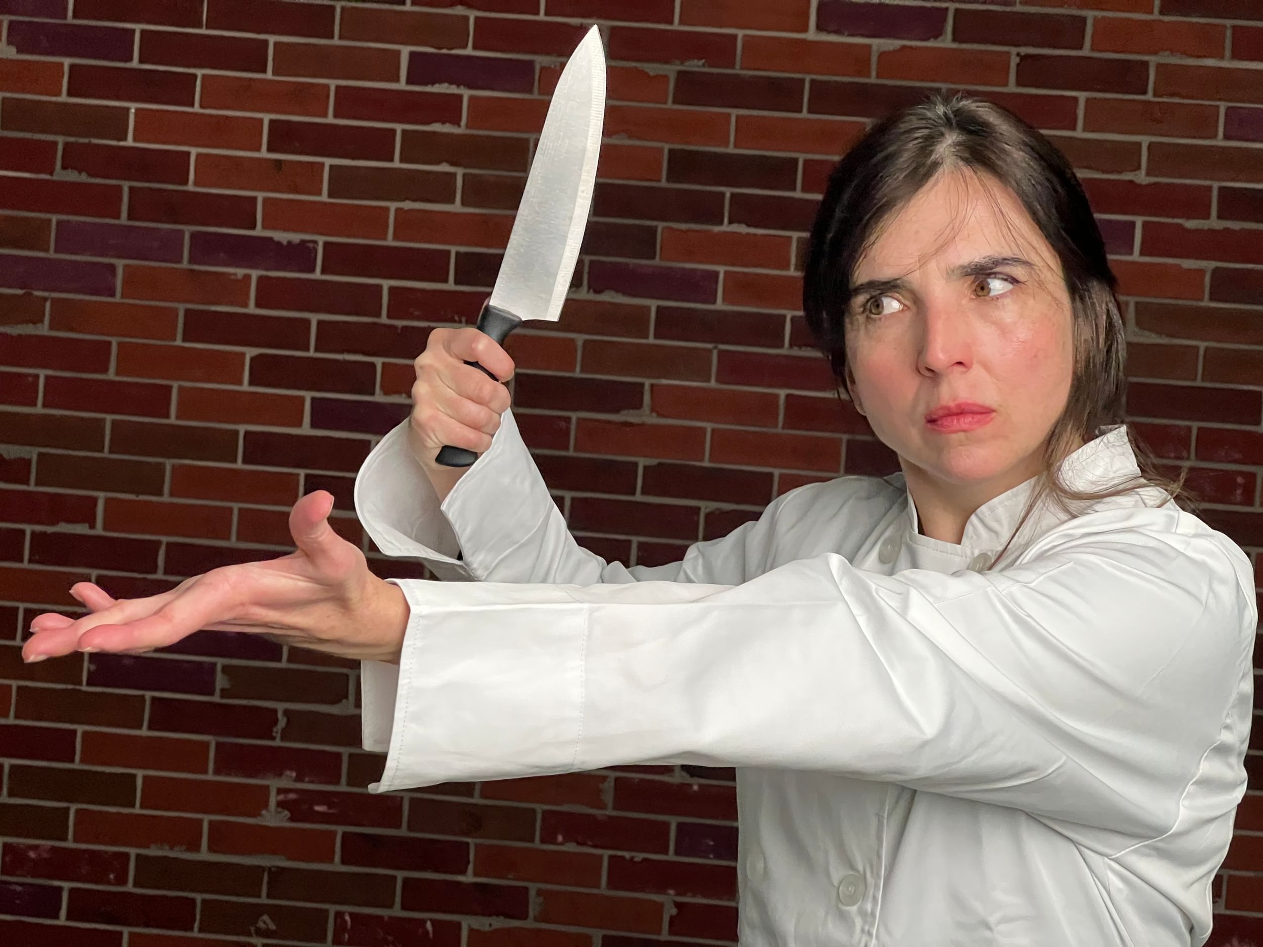 Allvin in a chef’s jacket, posits one arm extended out with fingers stretched, other hand holds a chef’s knife.