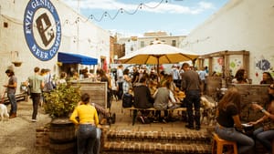 An outdoor beer garden is full with people, tables, parasols, light fixtures, and seating under a partly cloudy sky