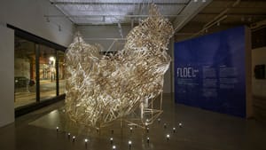 In a nighttime gallery room next to a large window, an airy, dramatically lit large sculpture of white bamboo strips.