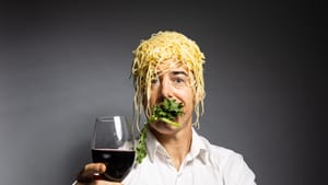 Sobelle holds a wineglass, toasting. Spaghetti glops on his head, his mouth full of greens. Fruits arranged in the foreground