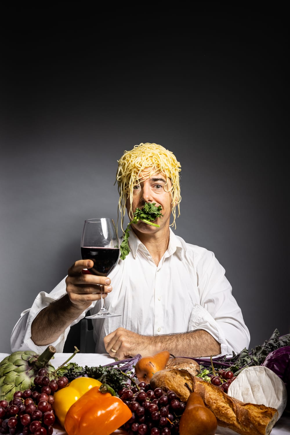 Sobelle holds a wineglass, toasting. Spaghetti glops on his head, his mouth full of greens. Fruits arranged in the foreground