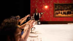 At left, a line of seated people raise wine glasses over a huge white table; Sobelle stands beyond them like a butler.