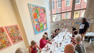 Seated at a long table full of arts supplies next to a large window, 14 people make colorful designs while Henbo watches.