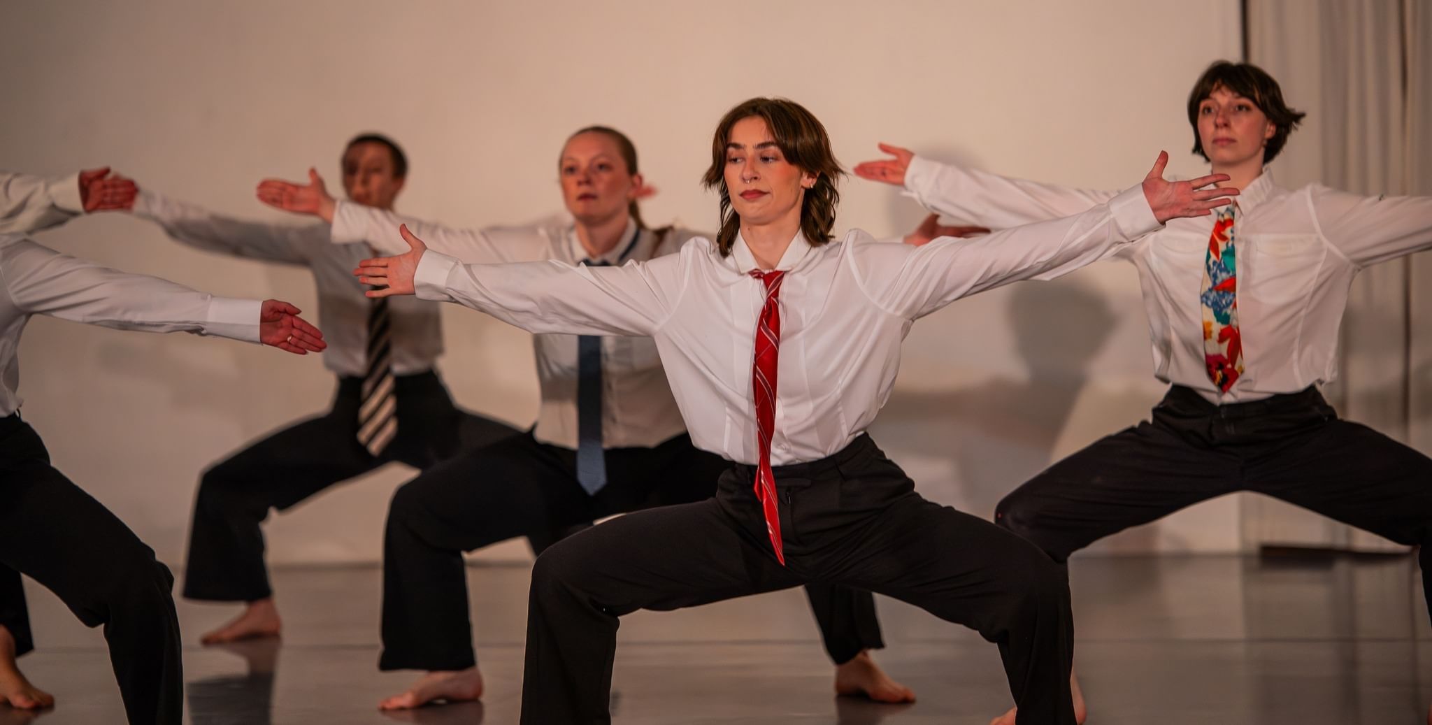 A group of dancers in white button down tops, ties, and black pants stretch out their arms and extend out their legs