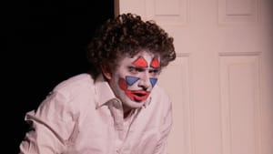 Spangler, in clown makeup, hunches over a bathroom sink. They have a curly mop of hair and wear a white button-down shirt