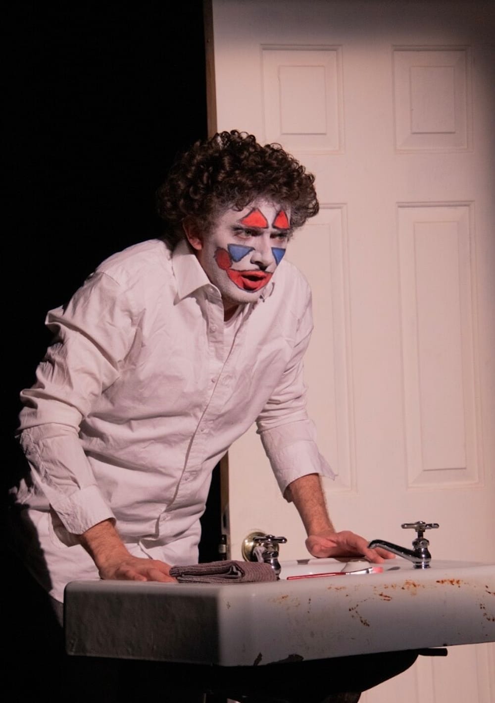 Spangler, in clown makeup, hunches over a bathroom sink. They have a curly mop of hair and wear a white button-down shirt