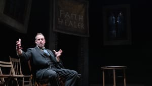 In a worn, dark suit, Peakes sits and gestures broadly in a wooden folding chair on a darkened stage of wooden planks.