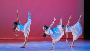 Three dancers in white dresses synced in mid-dance on a pink and purple lit stage