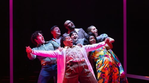 The 7 cast members stand close together, connected with arms and hands, leaning back together and singing.