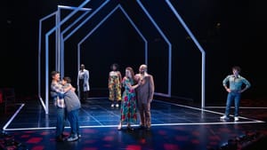 Within a stylized white stage outline of a house, a man in a plaid shirt hugs a kid while five other adults look on.