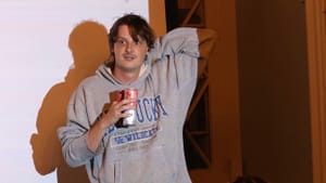 Giltner, a white person in a baggy gray sweatshirt, stands in front of a slideshow, holding a can of White Claw seltzer.