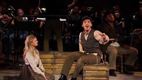 A woman watches a man singing out to the audience, the orchestra lined up in the background.