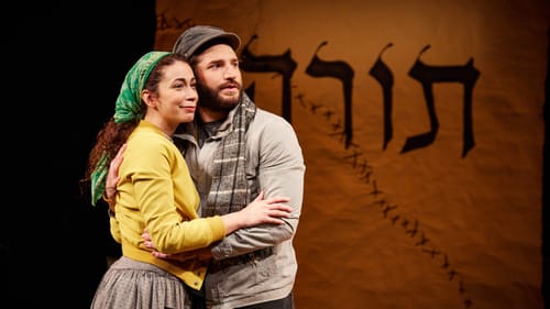 Lynne and Seigla embrace cheek to cheek, the banner with the Hebrew letters Torah behind them, its rip diagonally stitched.