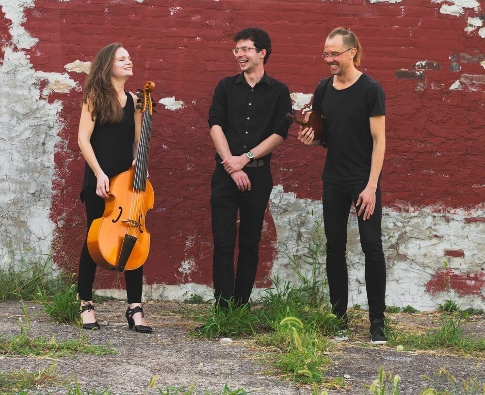 Three people in all black outdoors against a brick wall with decaying paint. One person holds a viola da gamba.