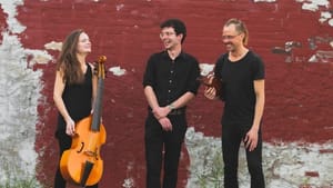 Three people in all black outdoors against a brick wall with decaying paint. One person holds a viola da gamba.