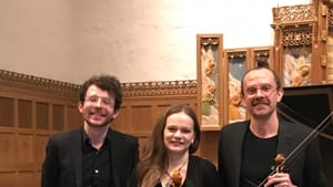 The three musicians stand together smiling on a small church chancel, Kaufman and Few holding their instruments.
