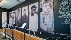 Six large b&w portrait photos of adults, one child, on tapestries at an exhibit wall with touch pads and headphones in front