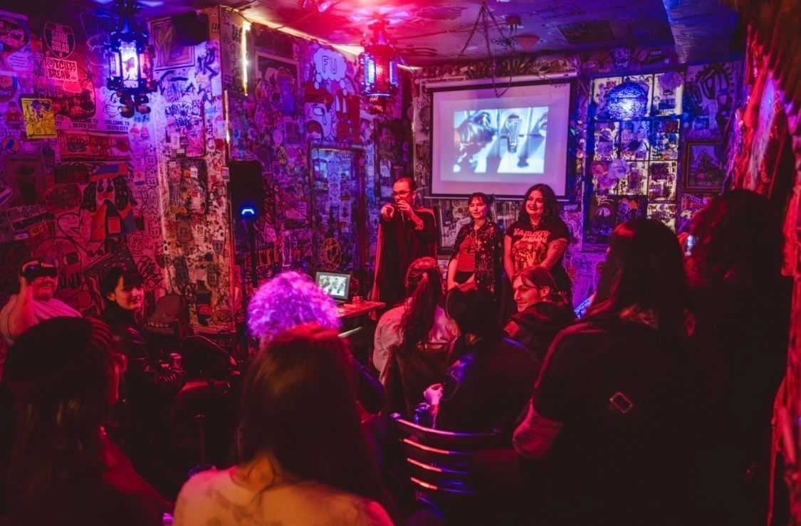 In a crowded space with neon light tones and walls covered in street art, three people stand on a small stage.