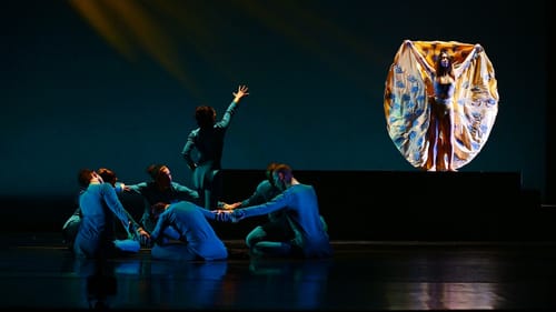 6 dancers in blue writhe in a circle on the floor; 1 reaches toward another who holds out a dramatic orange cape.