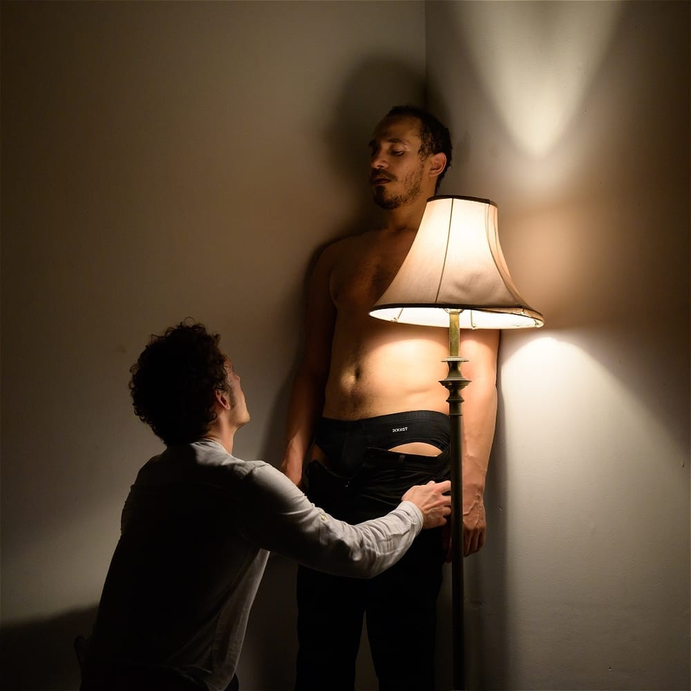 Grinberg, on his knees, faces the shirtless Header in a moment of sexual tension, next to a single lamp.