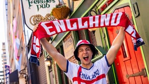 A person holds up an England scarf with excitement outside of a pub, wearing an England soccer jersey.