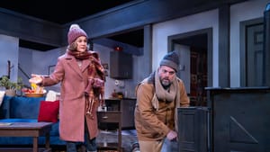 Childs & Greer, bundled up against the cold, argue in front of a woodstove in the large living room of a winter vacation home