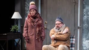 Childs stands next to Greer, who huddles in an armchair. They both wear thick hats, coats, and scarves indoors.