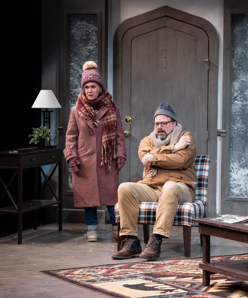 Childs stands next to Greer, who huddles in an armchair. They both wear thick hats, coats, and scarves indoors.