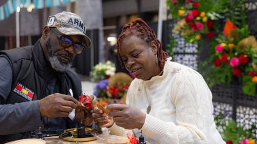 A Black man & woman lean intently over a small table with blossoms and craft supplies.