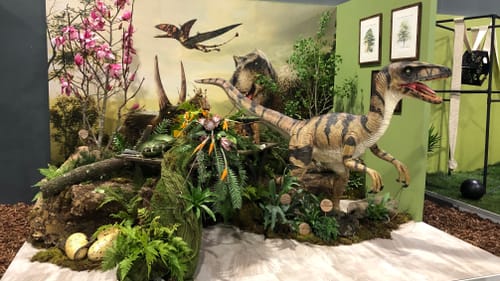 A prehistoric-looking display of logs, moss & ferns has a mural of dinosaurs behind it & a life-sized raptor stepping out