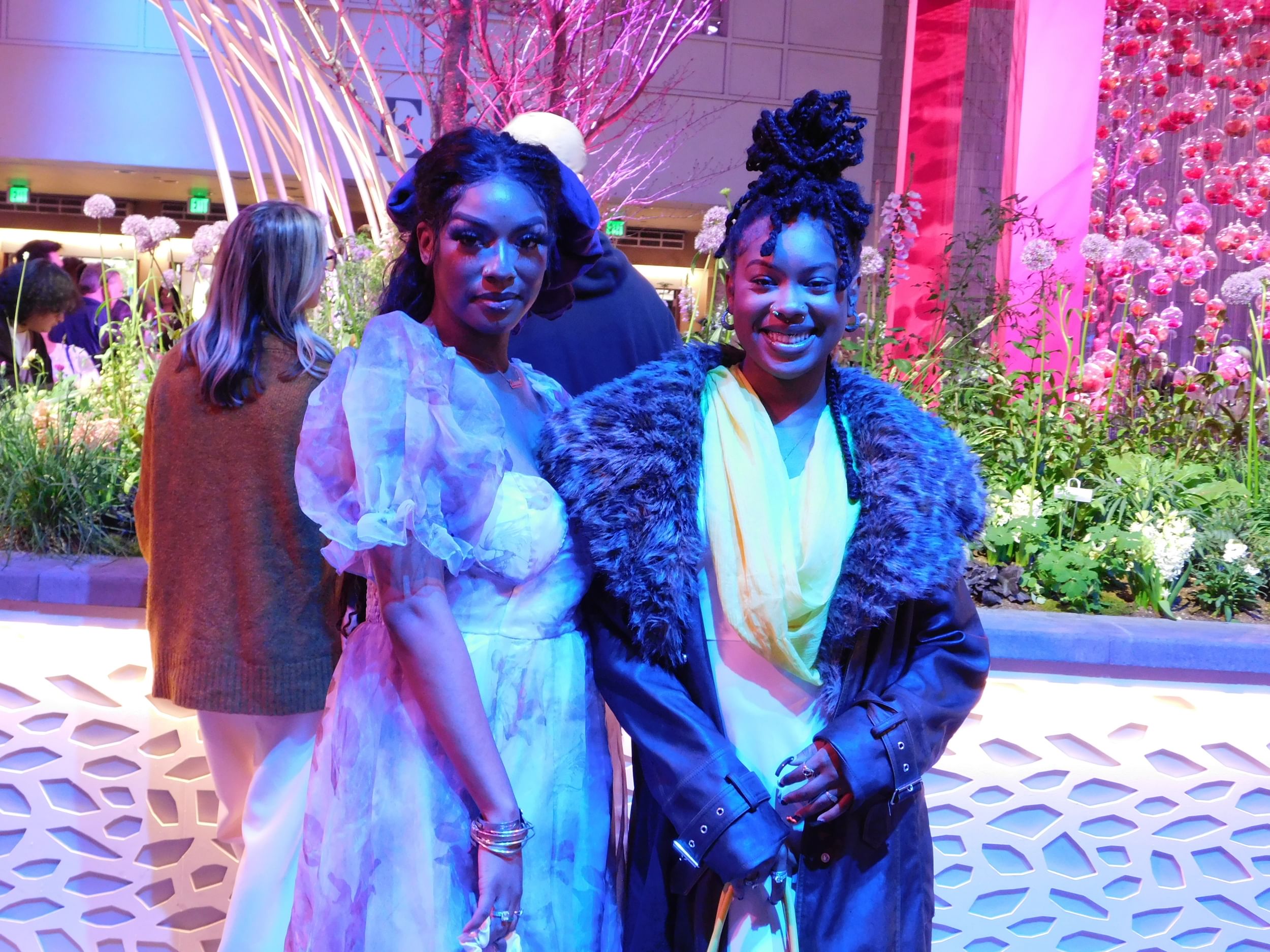 Two Black women pose smiling together in dresses with lavish color, shape, texture, and accessories.
