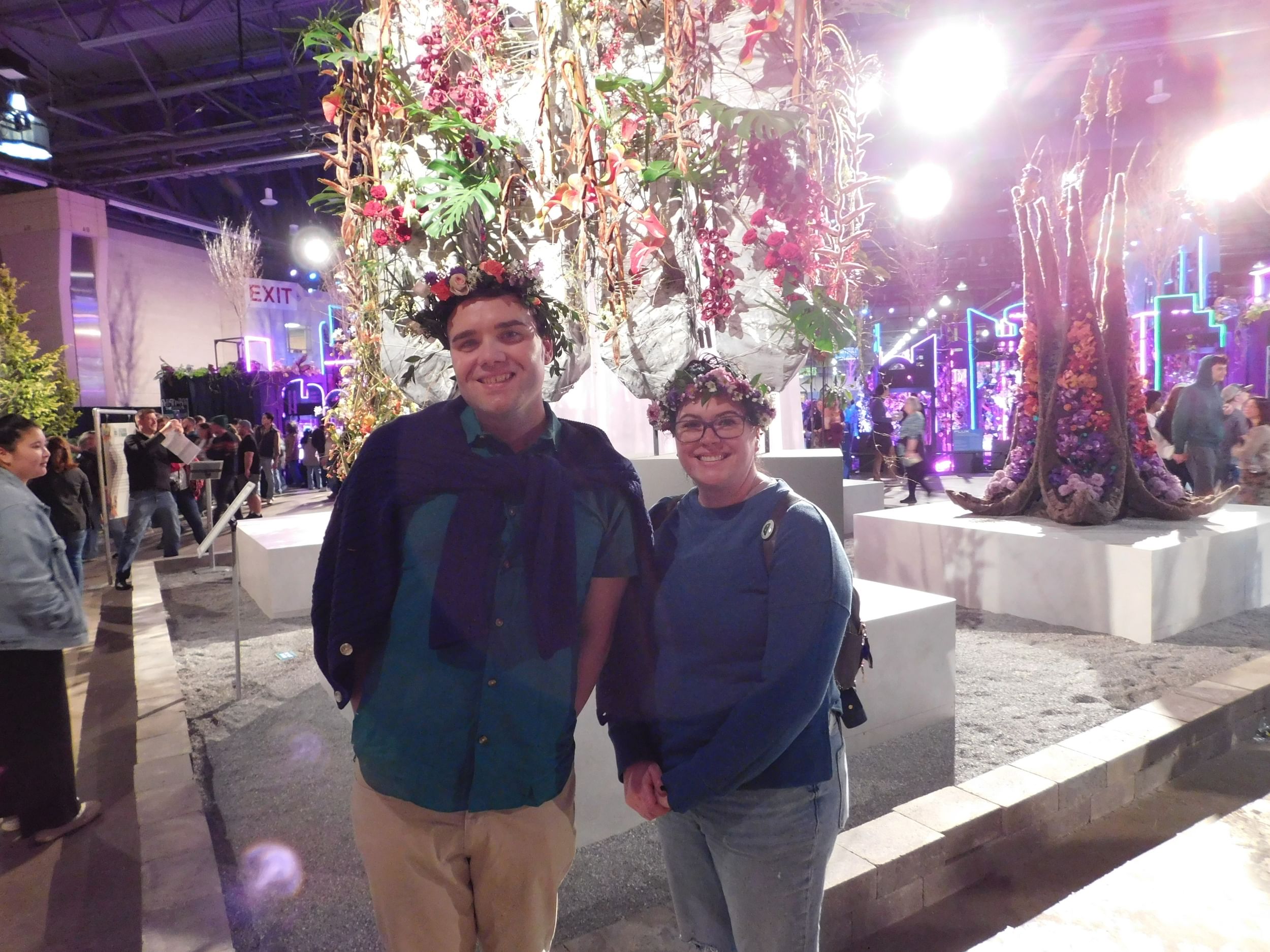 Two white people pose together smiling, wearing blue shirts and flower crowns.