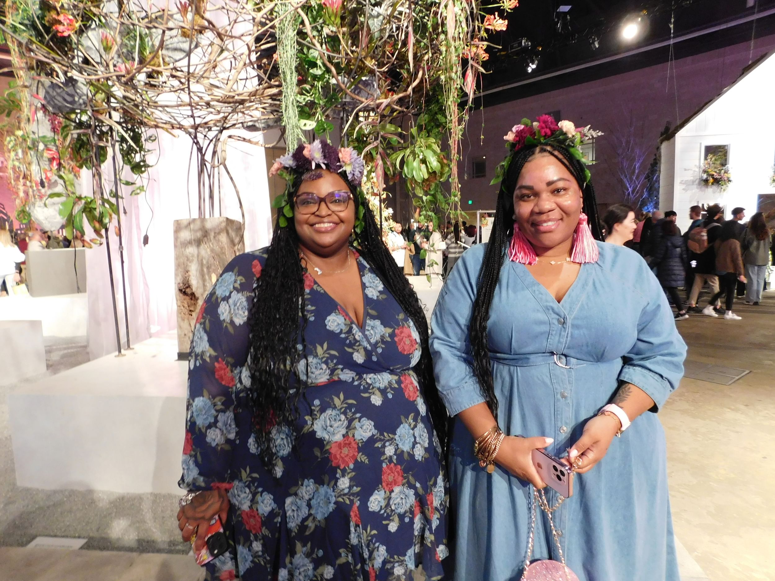 Two Black women wearing flower crowns smile together, one in a blue floral-patterned dress and the other in a sky-blue one.