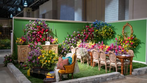 A green-walled life-sized diorama featuring a narrow table set for 12, practically disappearing under massive flower bouquets