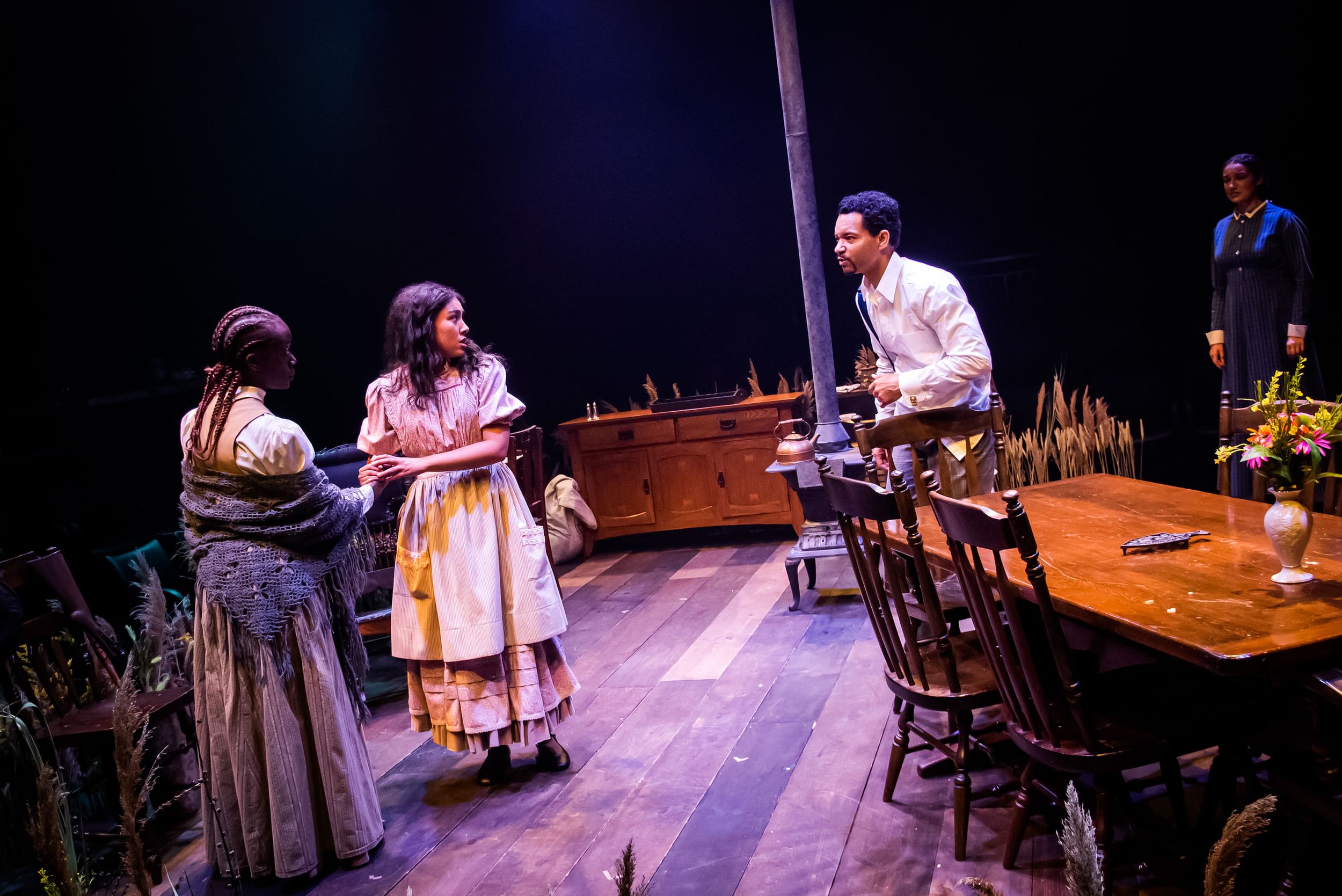 Four Black actors in period costume stand soberly in a late 19th-century interior, long grasses at the side, & black backdrop