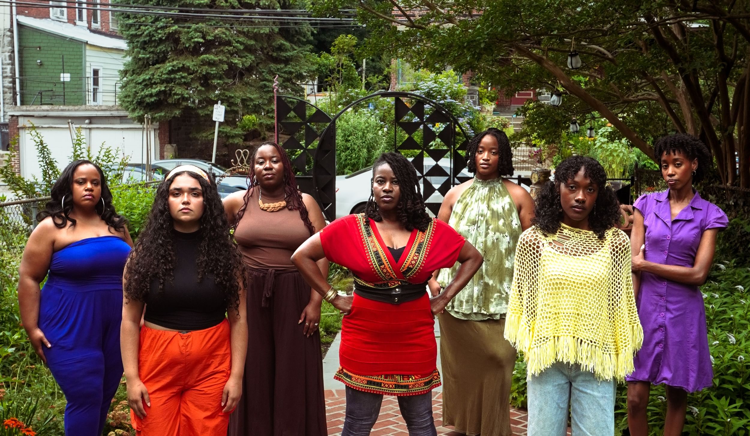 Seven Black women, each styled differently and wearing a different color, pose together with serious expressions in a park.