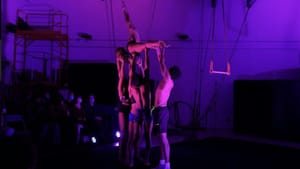 A group of dancers hold up another dancer in a dim space with deep purple lighting