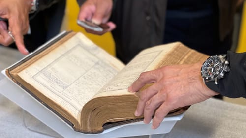 Close-up on the yellowed Bible, open about 4/5 of the way through. It’s a few inches thick and surrounded by gentle hands.