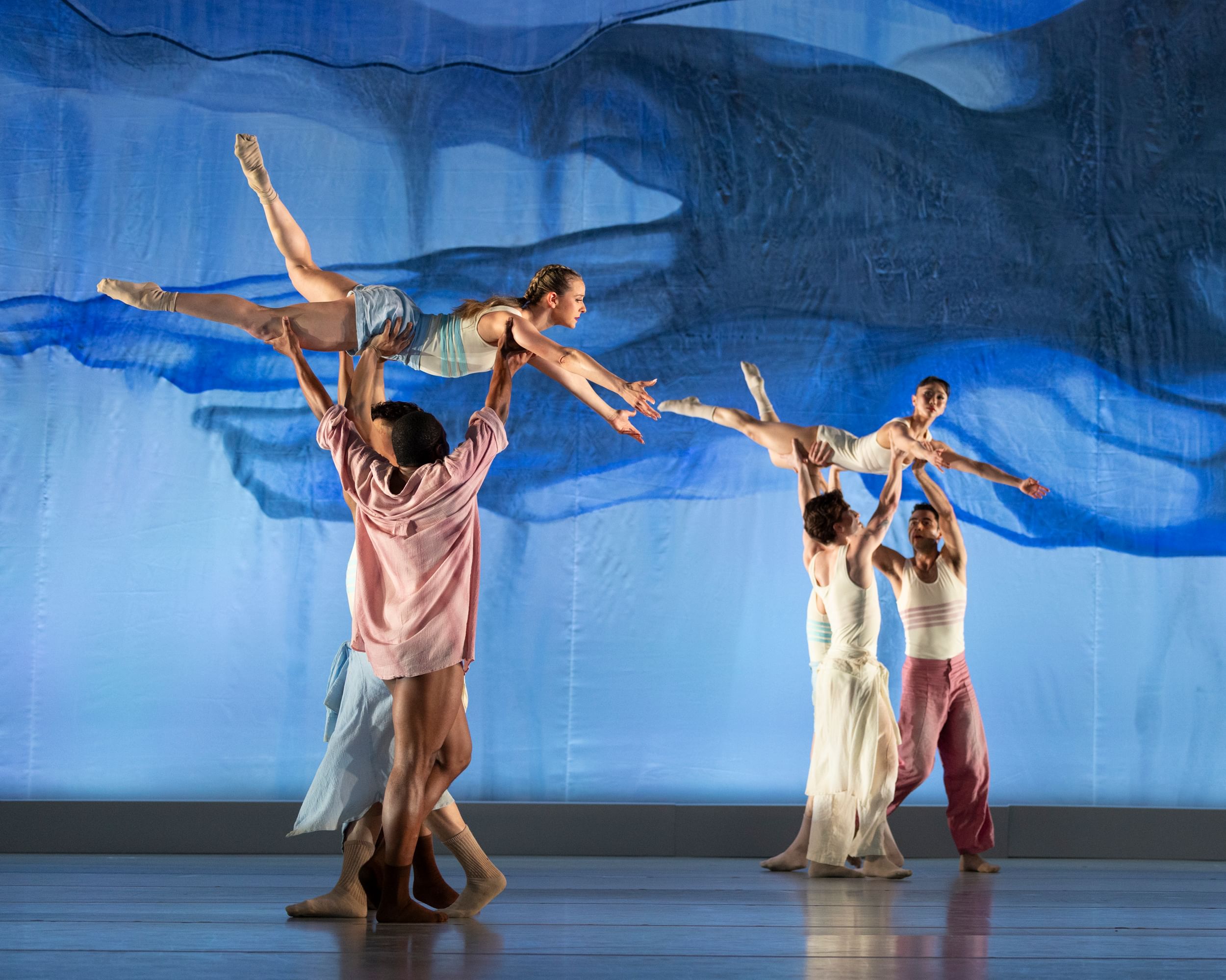 Two pairs of three dancers lift up another dancer on a stage, with blue lighting and a textured backdrop