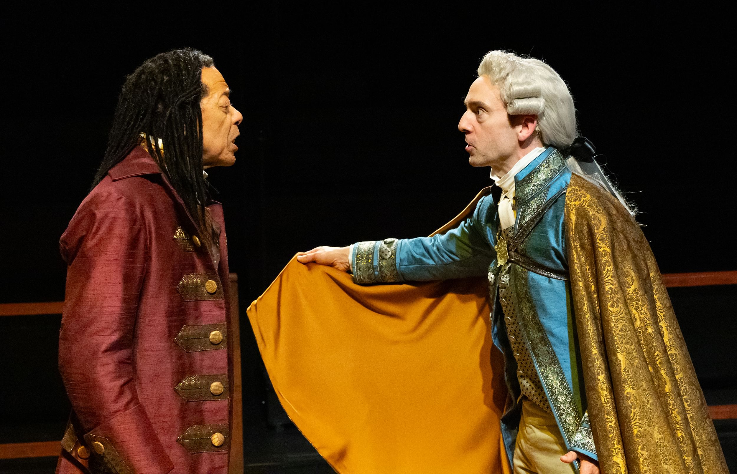 Closeup on X and Johnson facing each other as if they’re arguing, wearing opulent satin 18th-century costumes.