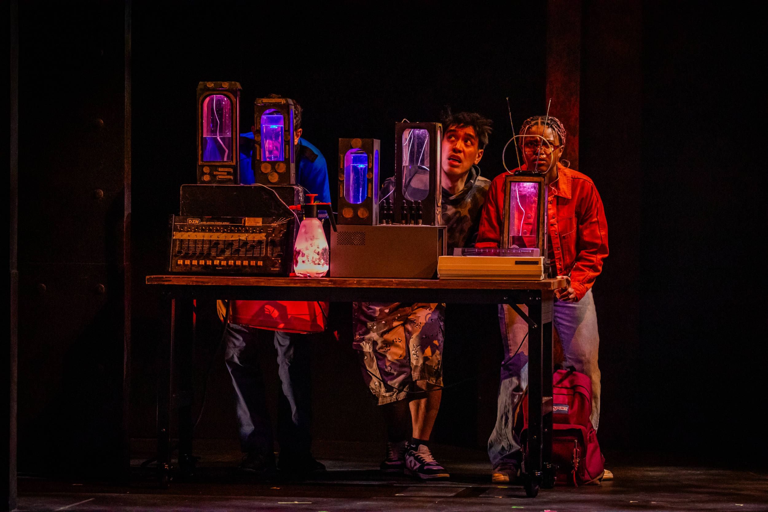 The three actors crouch worriedly behind a table with fantastical, neon-lit science gadgets.