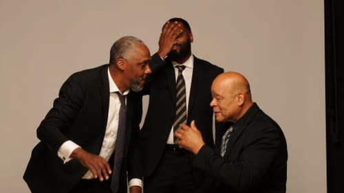 The three Black men, in dark suits, pose together as if gossiping. DeShields, at center, slaps a palm to his face.