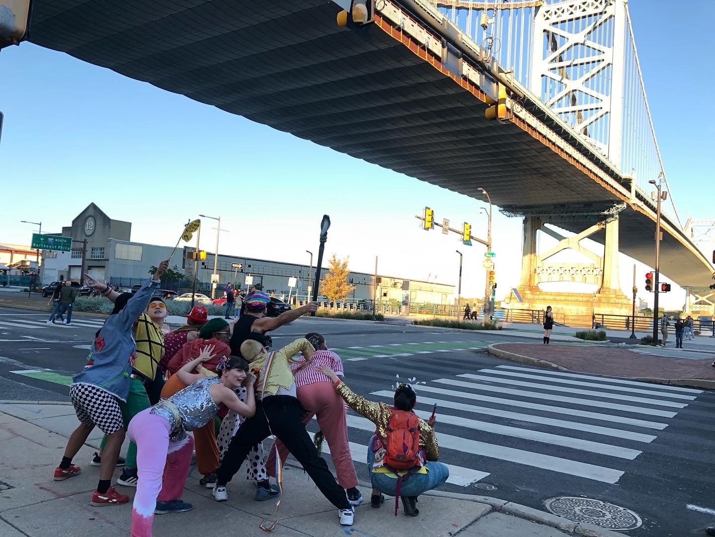 9 actors pose in a human sculpture at a crosswalk under a big bridge, hands touching each other's backs.
