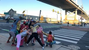 9 actors pose in a human sculpture at a crosswalk under a big bridge, hands touching each other's backs.