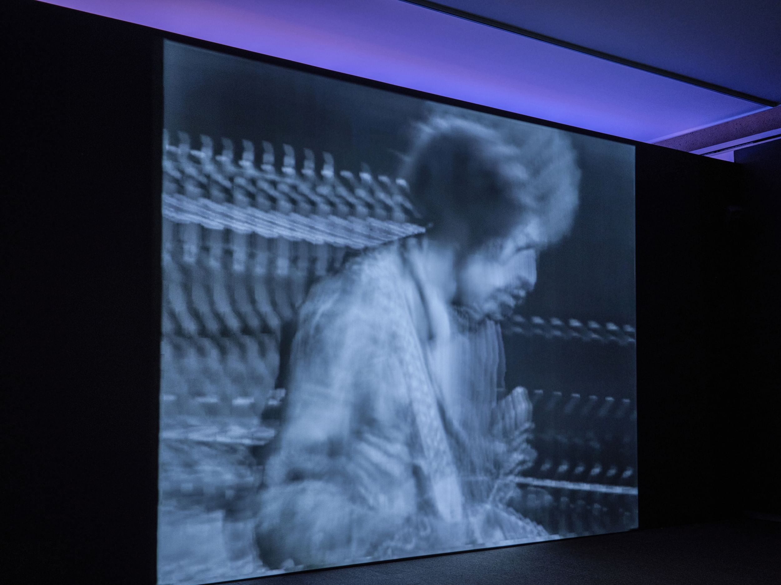 In a dark gallery, a stylishly blurred image of Jimi Hendrix is projected on a screen below a bar of blue in the ceiling.