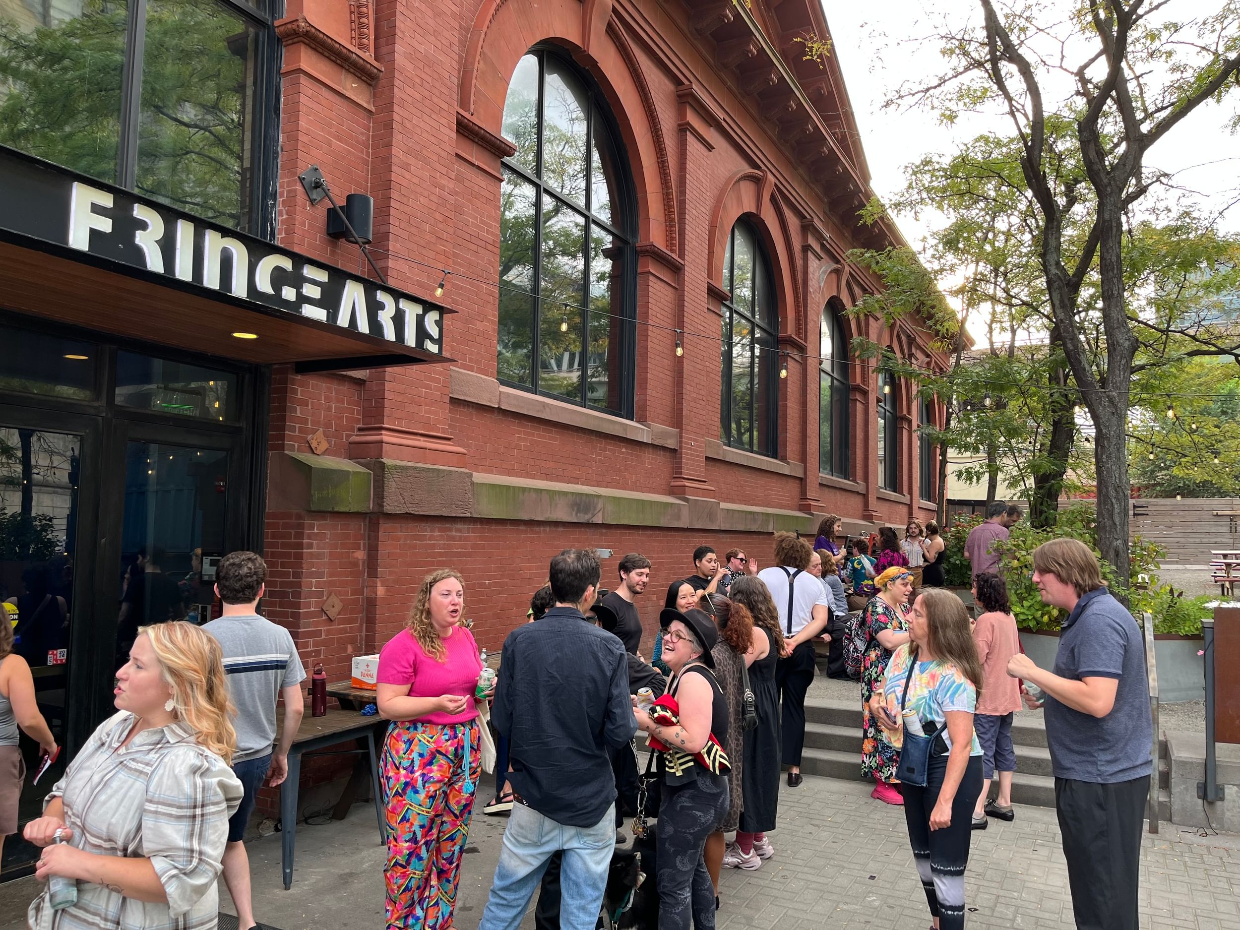 A crowd of people in different colorful clothes socialize outside the tall brick façade of the FringeArts building.