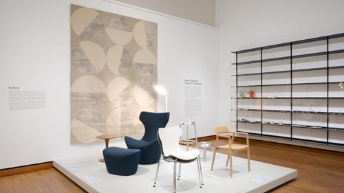 5 different stools and chairs, including pieces described above, sit on a low white rectangular dais in the gallery