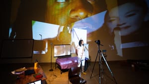 Gaines in front of a camera on a tripod, a bed behind him, and a collage of images projected at back of stage