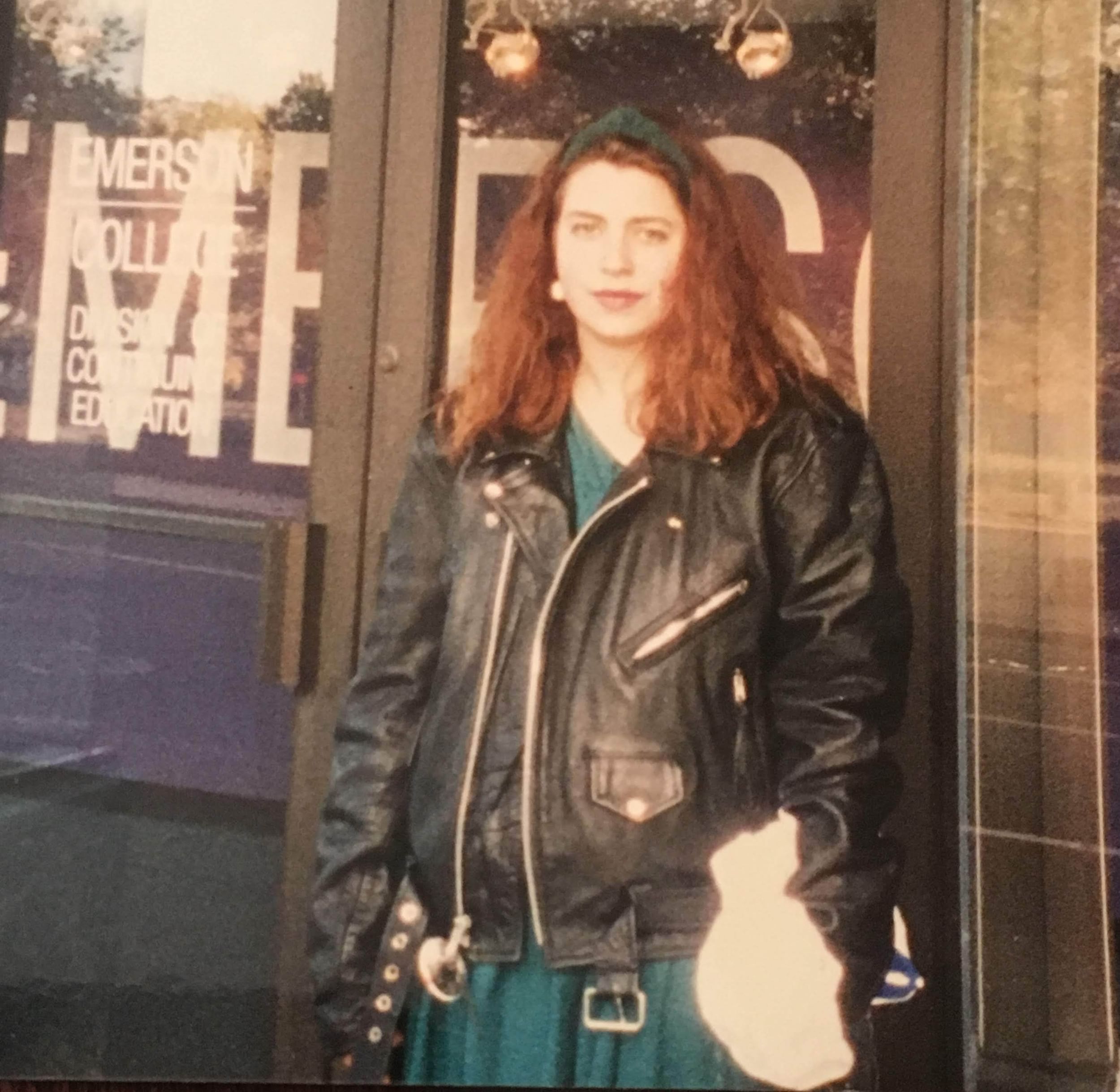 Gabrielle leans against a storefront. She wears a black leather jacket, green headband, and has tousled reddish hair.