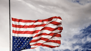 A large American flag flies upside down on a flagpole in the wind, against a cloudy sky.